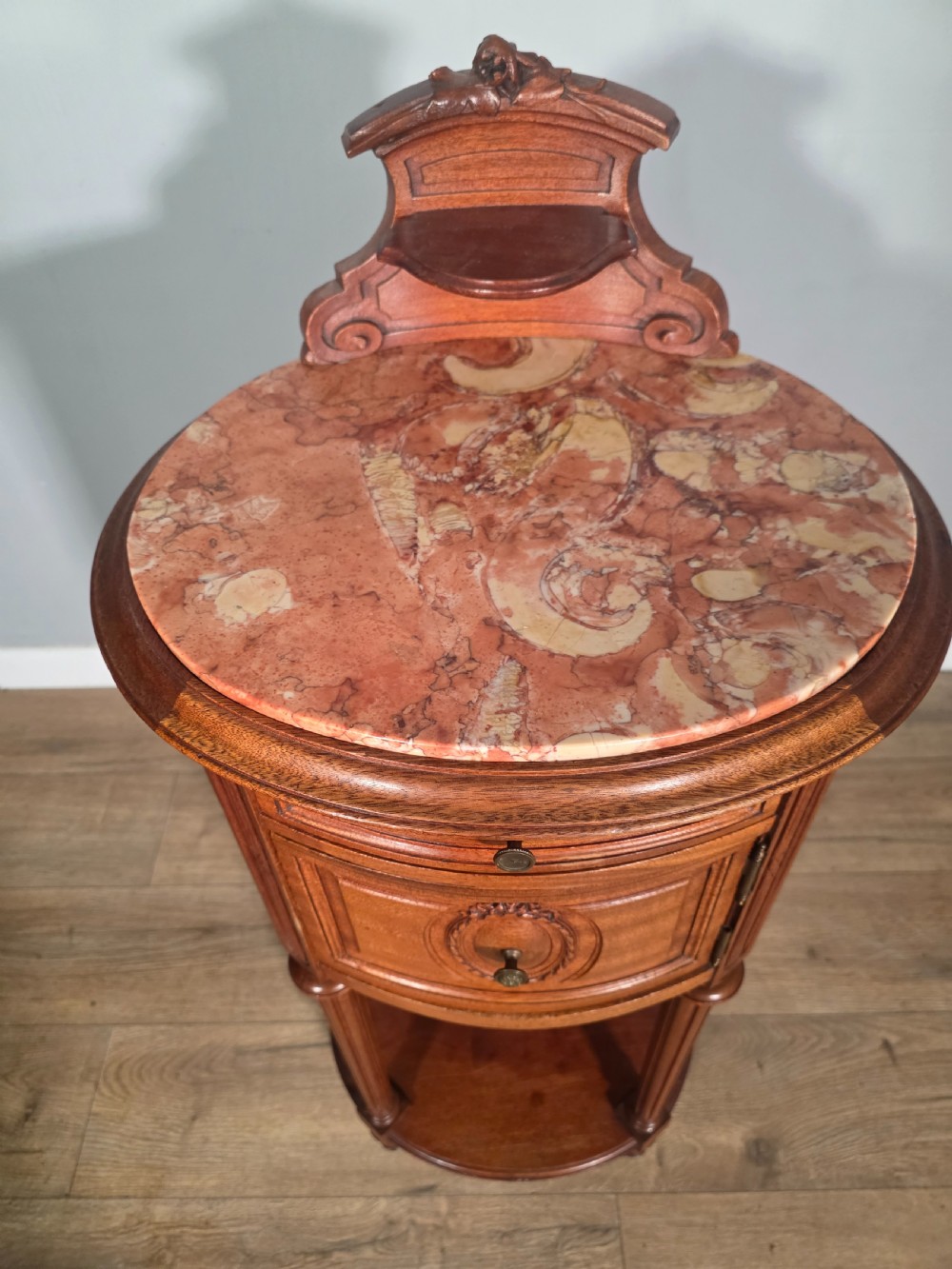 An antique French mahogany bedside cabinet featuring an oval marble top, a carved cresting gallery, a single drawer above a cupboard door, turned fluted legs, and a lower shelf. View IX