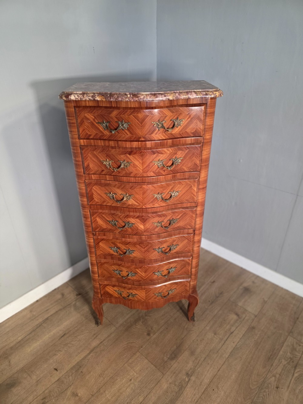 A tall 1930s French semainier chest featuring seven inlaid wood drawers, cast brass handles, a fitted brown marble top, and curved legs.
