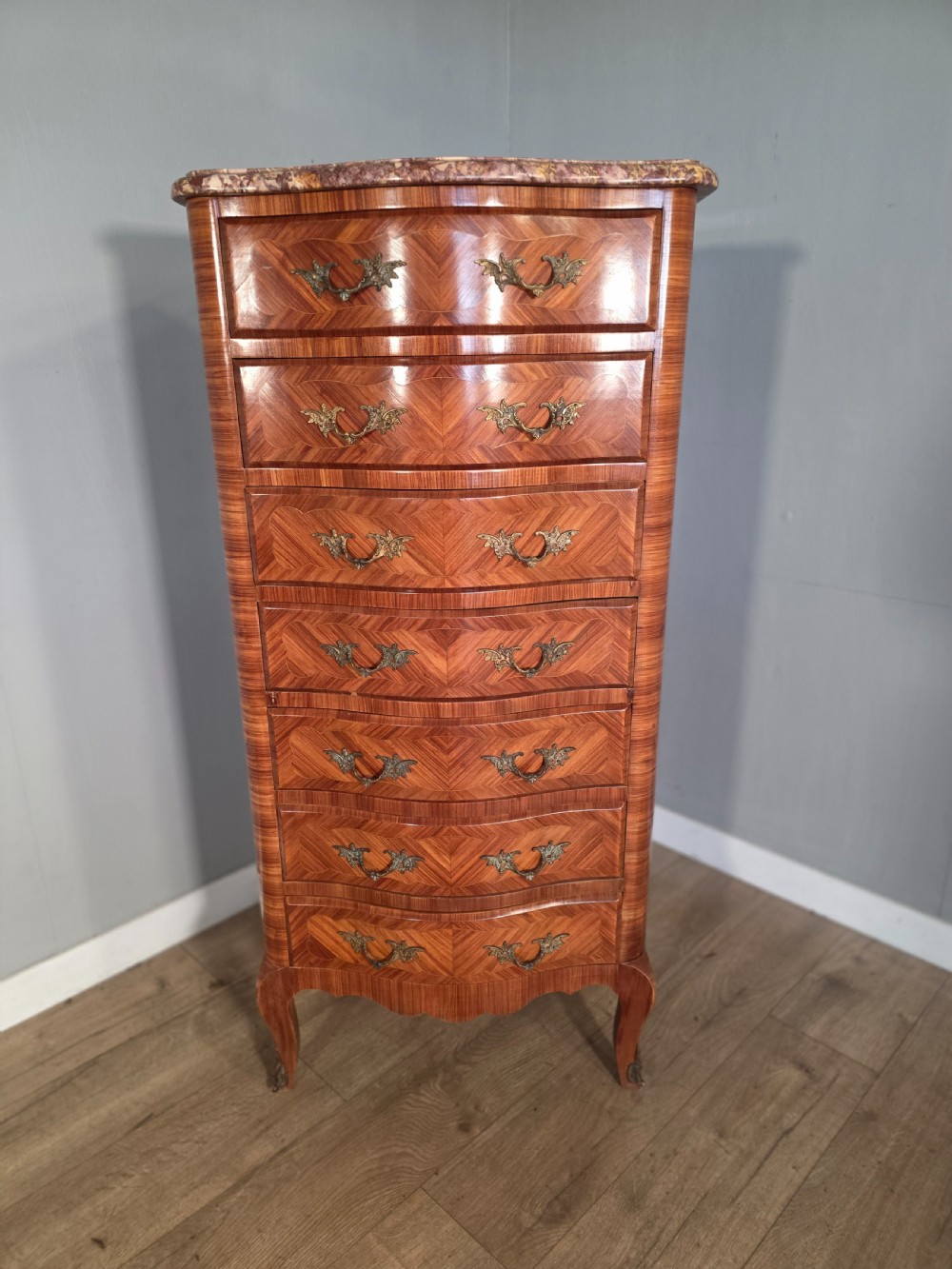 A tall 1930s French semainier chest featuring seven inlaid wood drawers, cast brass handles, a fitted brown marble top, and curved legs. View II