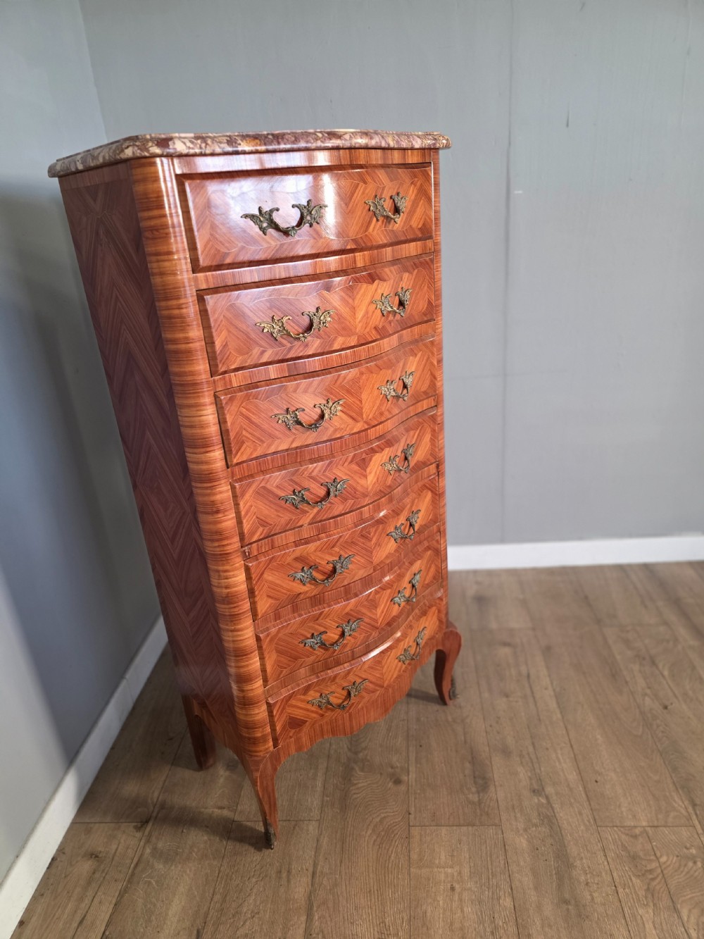 A tall 1930s French semainier chest featuring seven inlaid wood drawers, cast brass handles, a fitted brown marble top, and curved legs. View III