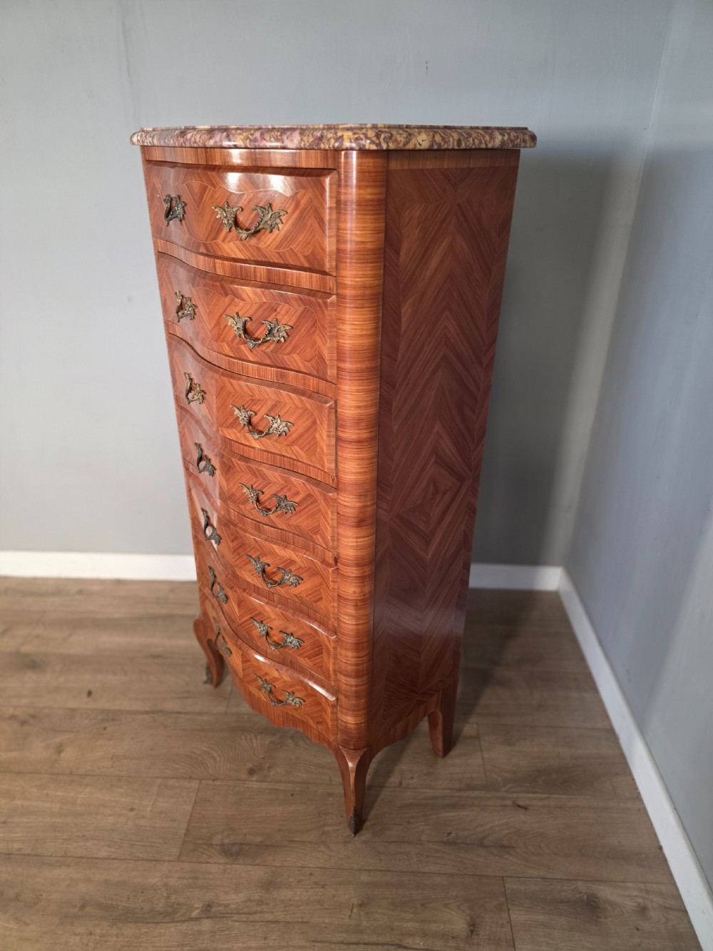 A tall 1930s French semainier chest featuring seven inlaid wood drawers, cast brass handles, a fitted brown marble top, and curved legs. View IV
