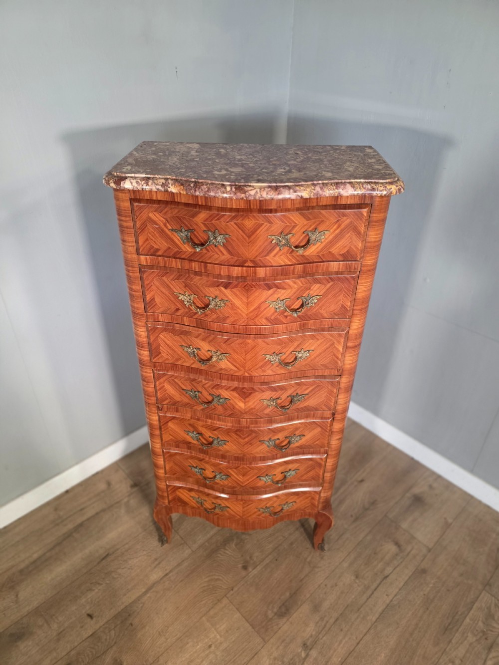A tall 1930s French semainier chest featuring seven inlaid wood drawers, cast brass handles, a fitted brown marble top, and curved legs. View V