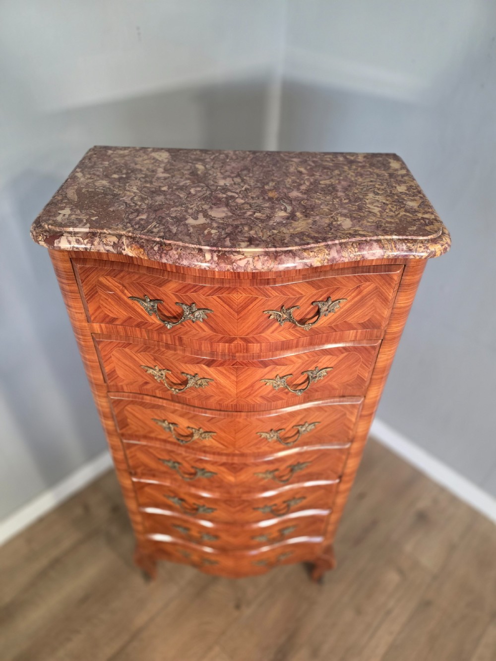 A tall 1930s French semainier chest featuring seven inlaid wood drawers, cast brass handles, a fitted brown marble top, and curved legs. View VI