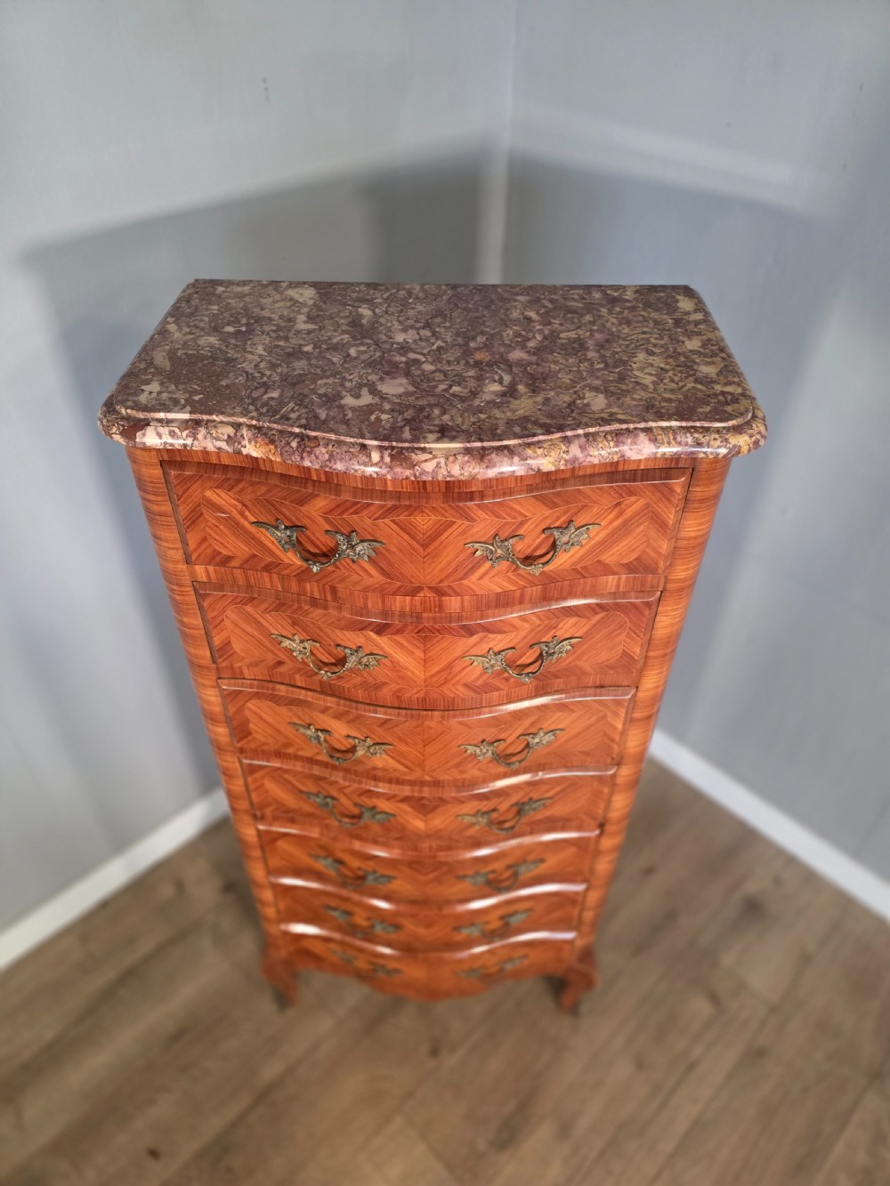 A tall 1930s French semainier chest featuring seven inlaid wood drawers, cast brass handles, a fitted brown marble top, and curved legs. View VII