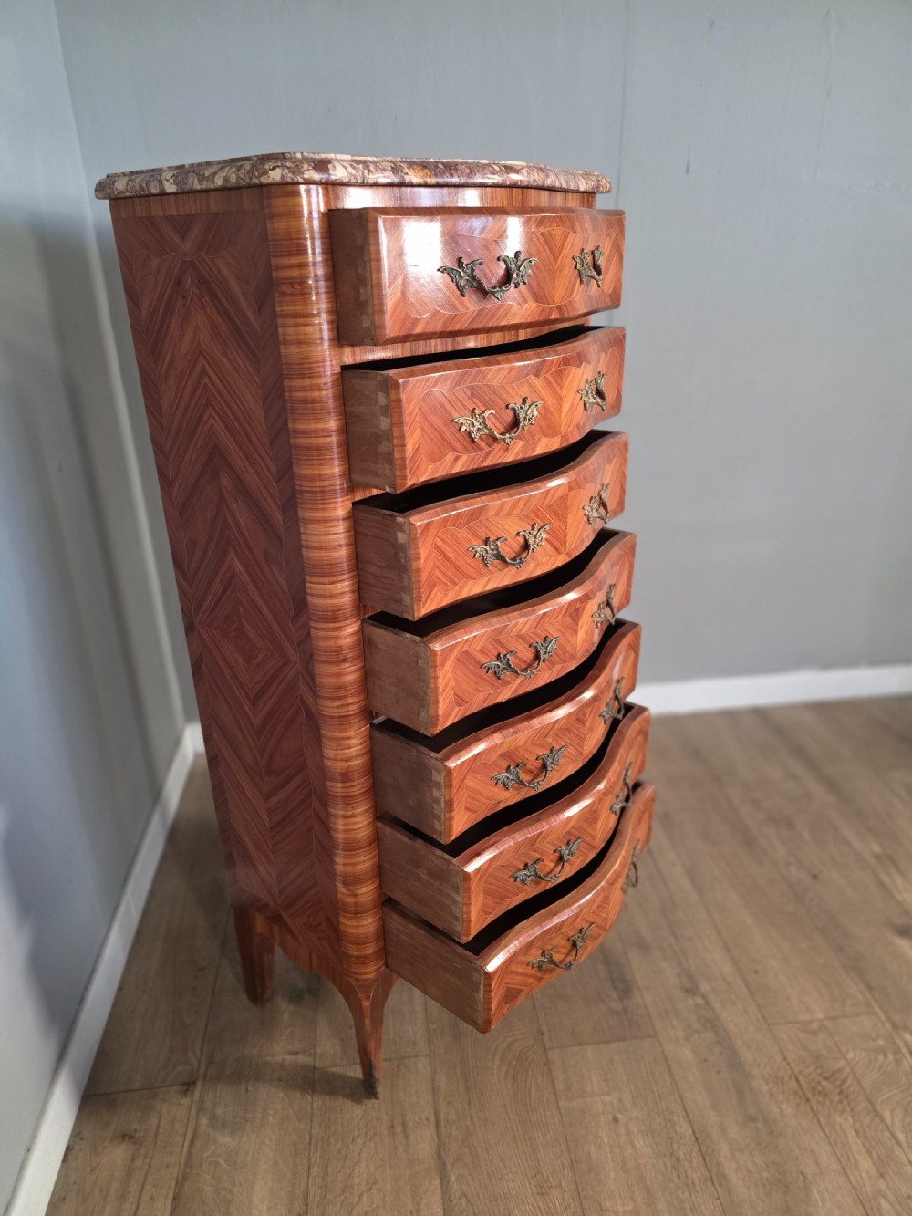 A tall 1930s French semainier chest featuring seven inlaid wood drawers, cast brass handles, a fitted brown marble top, and curved legs. View VIII