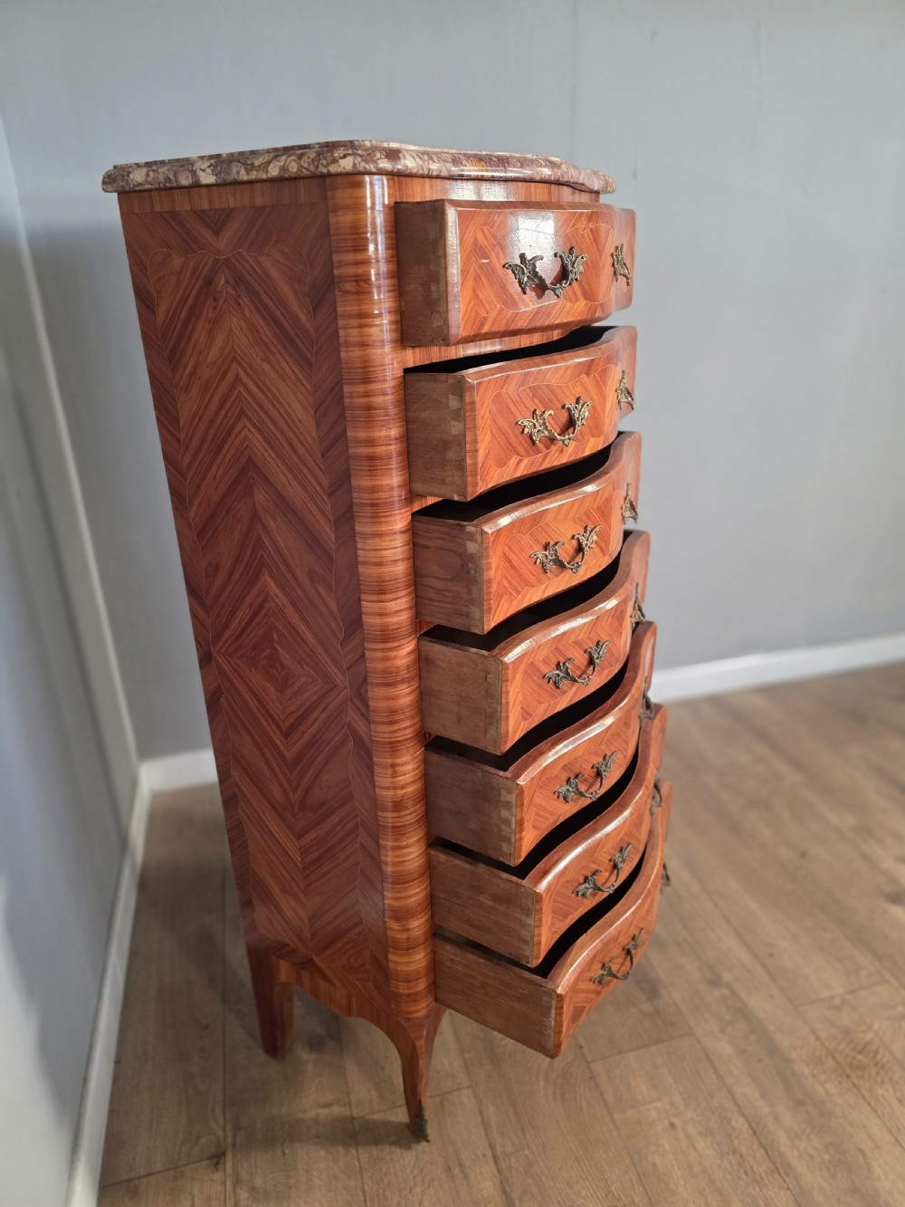 A tall 1930s French semainier chest featuring seven inlaid wood drawers, cast brass handles, a fitted brown marble top, and curved legs. View IX