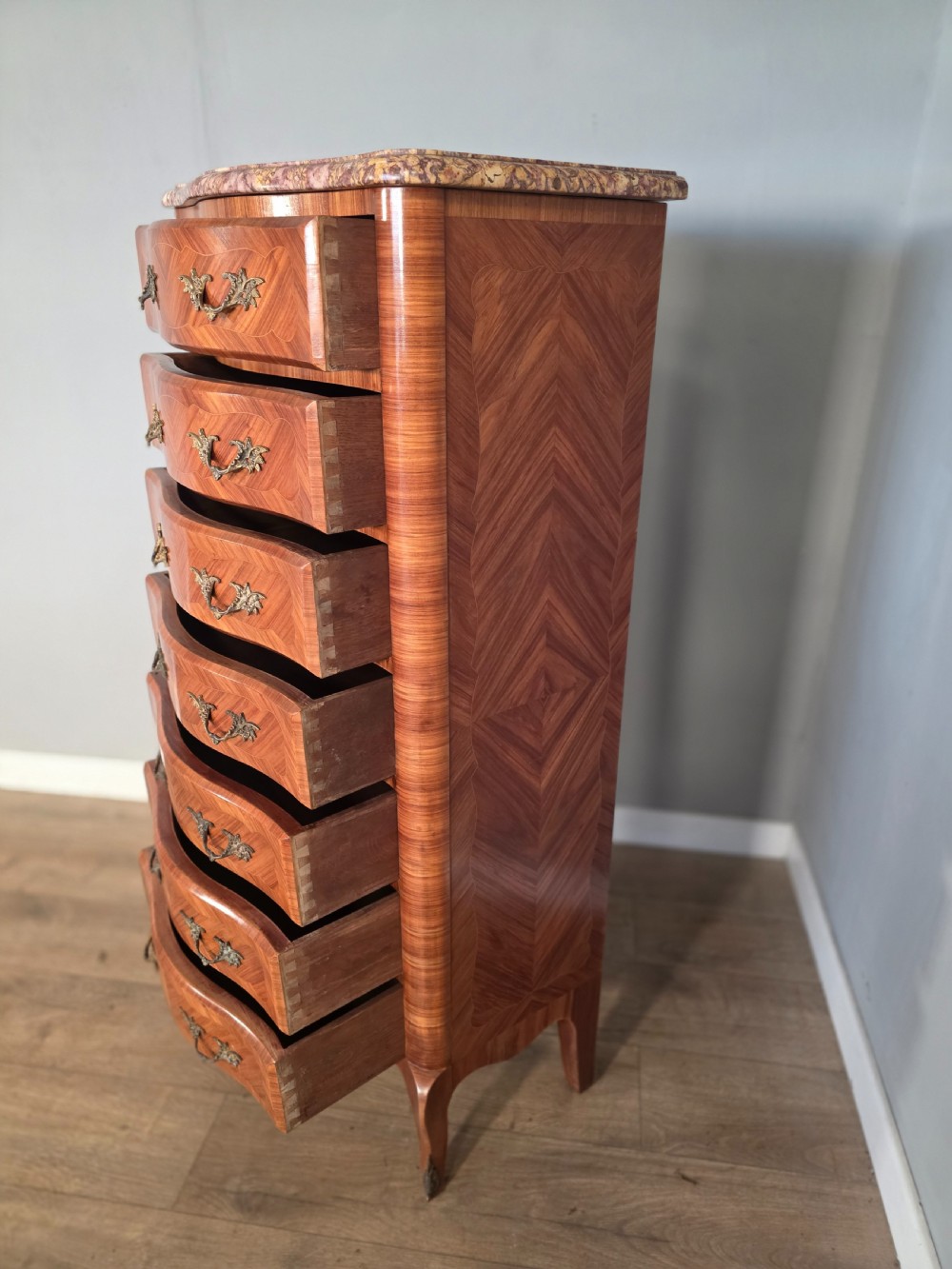 A tall 1930s French semainier chest featuring seven inlaid wood drawers, cast brass handles, a fitted brown marble top, and curved legs. View X