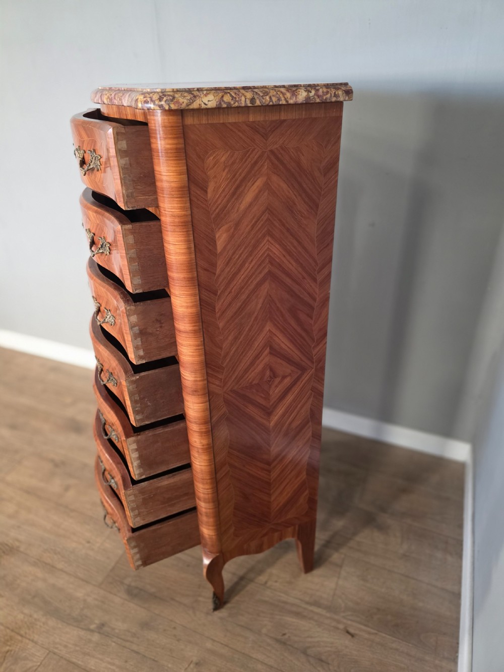 A tall 1930s French semainier chest featuring seven inlaid wood drawers, cast brass handles, a fitted brown marble top, and curved legs. View XI