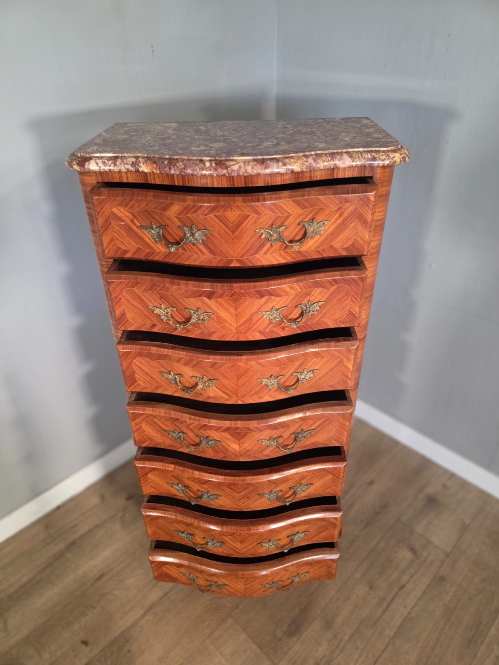 A tall 1930s French semainier chest featuring seven inlaid wood drawers, cast brass handles, a fitted brown marble top, and curved legs. View XII
