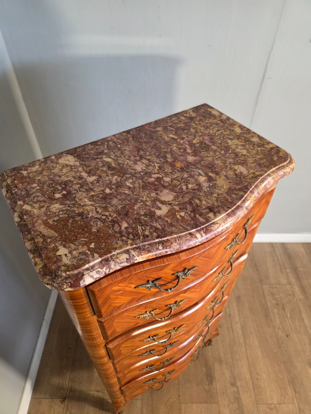 A tall 1930s French semainier chest featuring seven inlaid wood drawers, cast brass handles, a fitted brown marble top, and curved legs. View XV