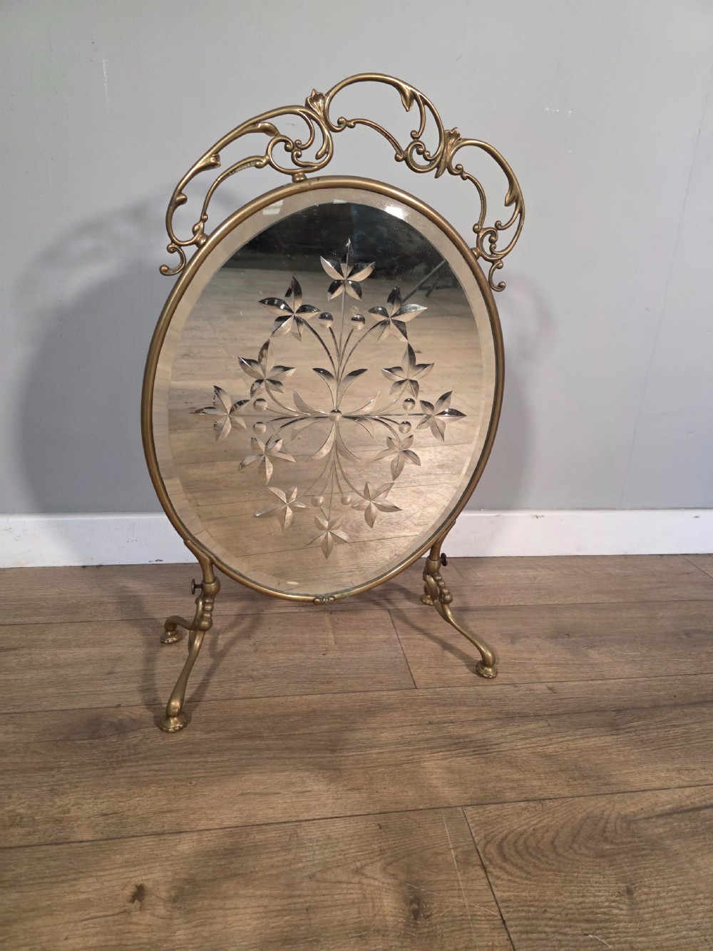 An Art Nouveau brass fire screen containing an oval bevelled glass panel engraved with a floral motif, set on splayed brass legs.