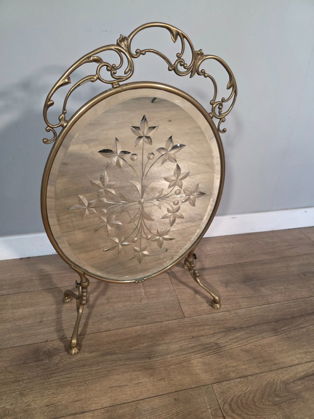 An Art Nouveau brass fire screen containing an oval bevelled glass panel engraved with a floral motif, set on splayed brass legs. View II