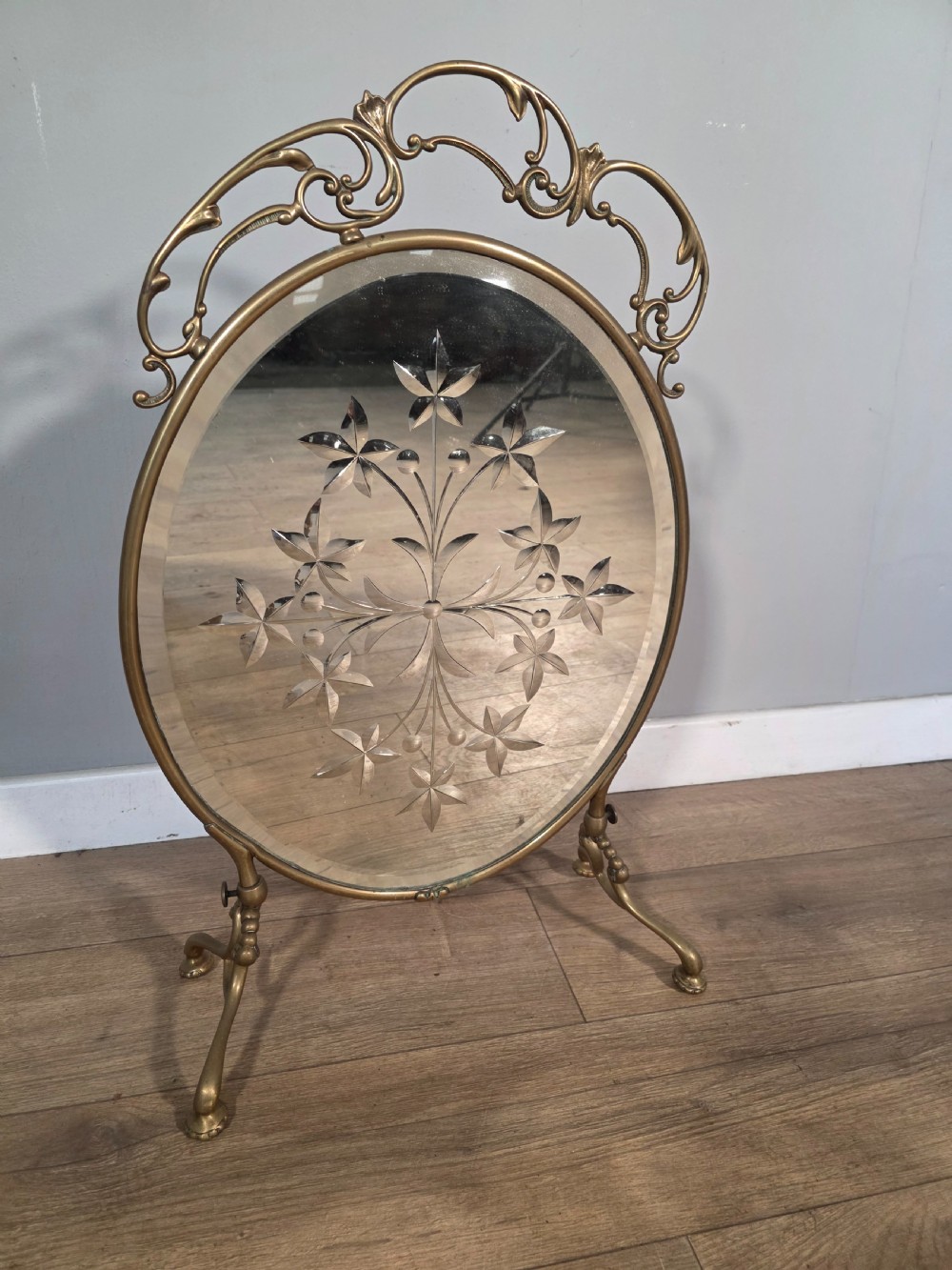 An Art Nouveau brass fire screen containing an oval bevelled glass panel engraved with a floral motif, set on splayed brass legs. View III