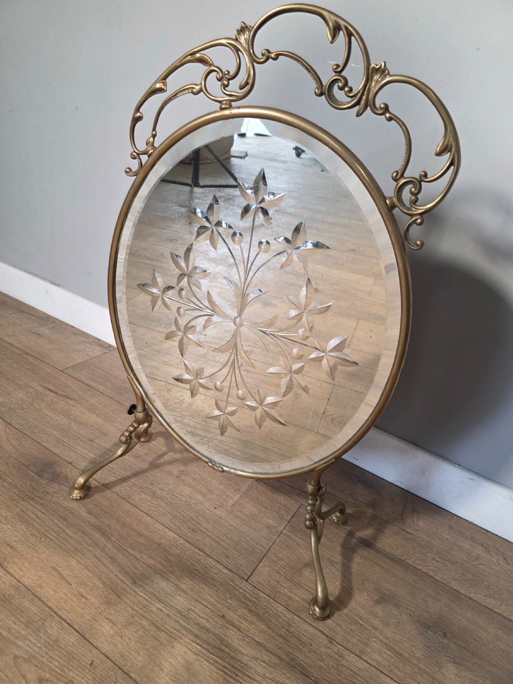 An Art Nouveau brass fire screen containing an oval bevelled glass panel engraved with a floral motif, set on splayed brass legs. View IV