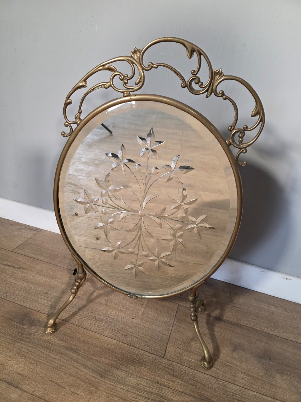 An Art Nouveau brass fire screen containing an oval bevelled glass panel engraved with a floral motif, set on splayed brass legs. View V