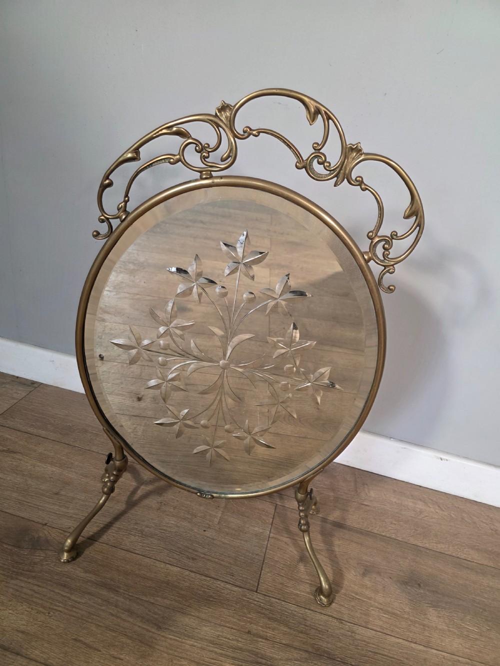 An Art Nouveau brass fire screen containing an oval bevelled glass panel engraved with a floral motif, set on splayed brass legs. View VI