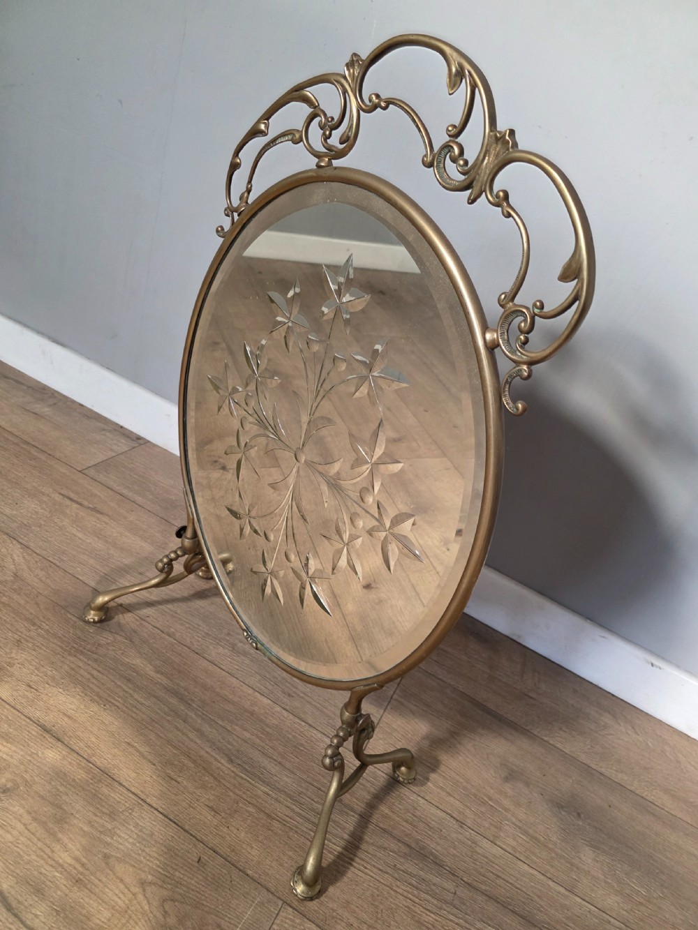 An Art Nouveau brass fire screen containing an oval bevelled glass panel engraved with a floral motif, set on splayed brass legs. View VIII