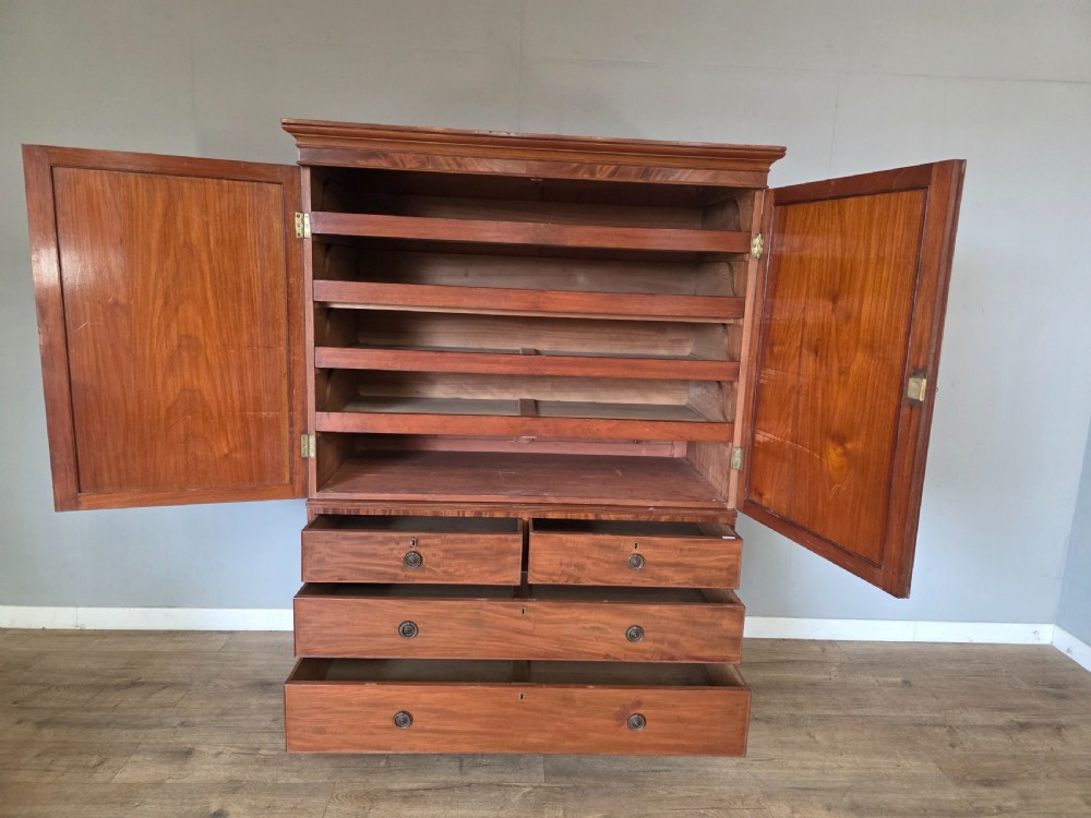 A George III mahogany linen press featuring flame mahogany panelled cupboard doors above a base of four drawers. View IX