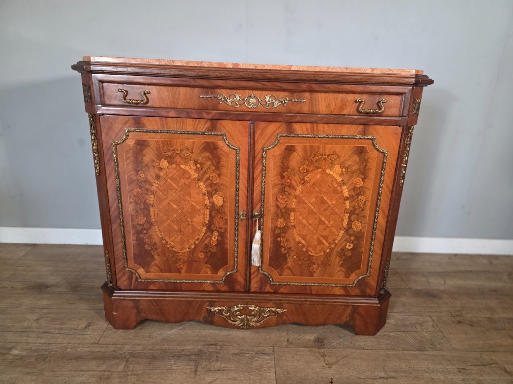 A 1920s French wooden cupboard featuring intricate floral marquetry inlay, gilt metal mounts, a single top drawer, two cabinet doors, and a reddish marble top surface.