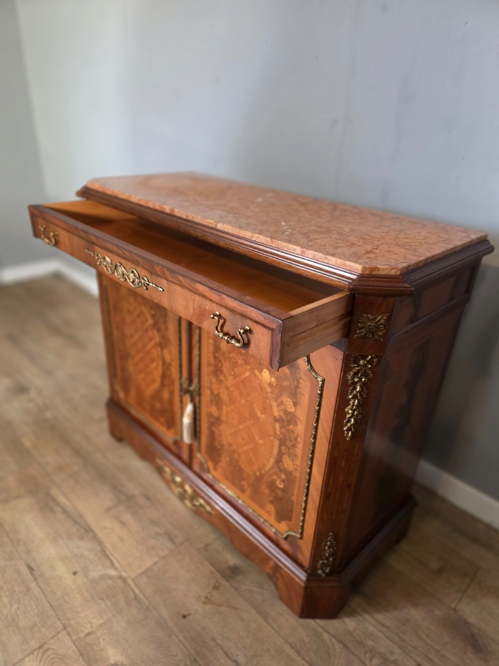 A 1920s French wooden cupboard featuring intricate floral marquetry inlay, gilt metal mounts, a single top drawer, two cabinet doors, and a reddish marble top surface. View IX