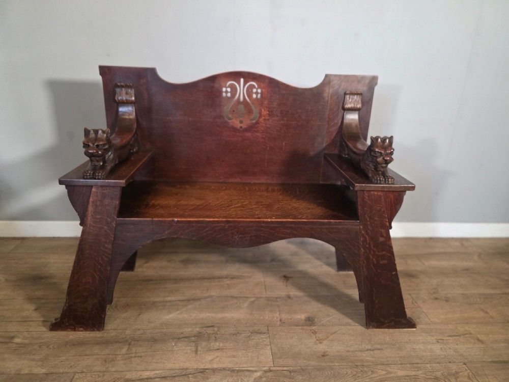 A dark oak Arts and Crafts hall bench featuring carved lion armrests, angled side legs, and a stylized inlaid motif on the backrest.