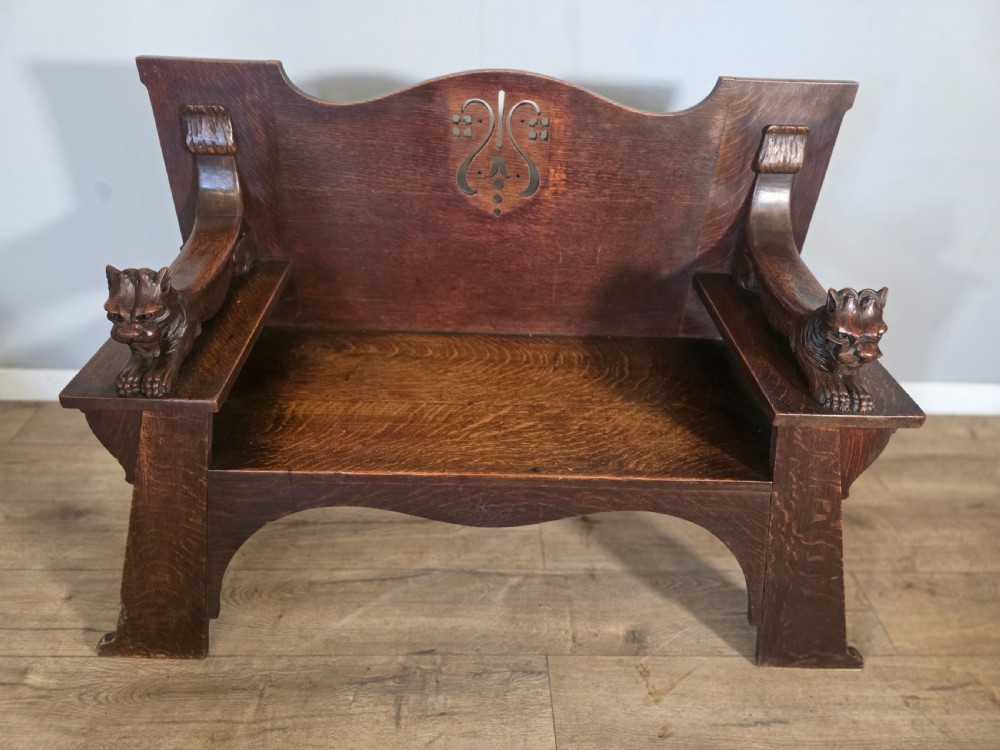 A dark oak Arts and Crafts hall bench featuring carved lion armrests, angled side legs, and a stylized inlaid motif on the backrest. View IV