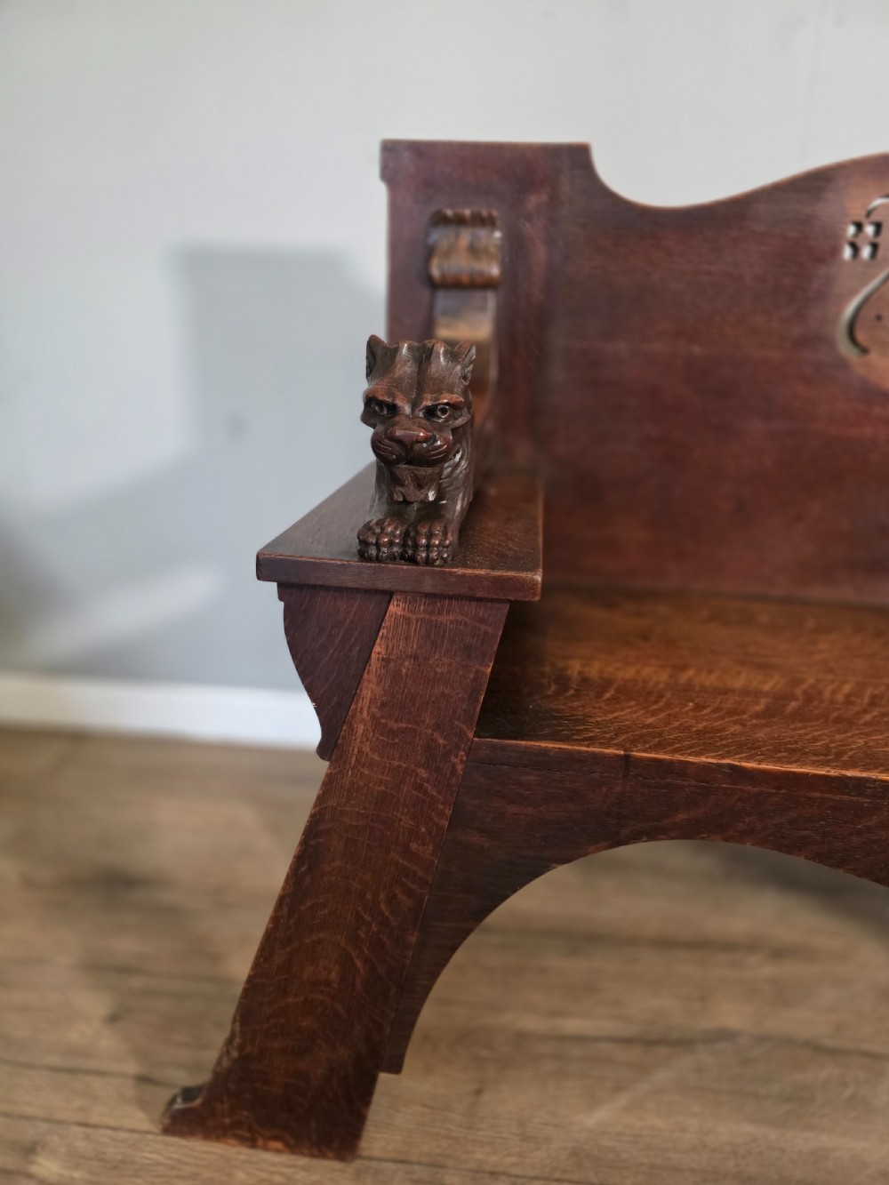A dark oak Arts and Crafts hall bench featuring carved lion armrests, angled side legs, and a stylized inlaid motif on the backrest. View V