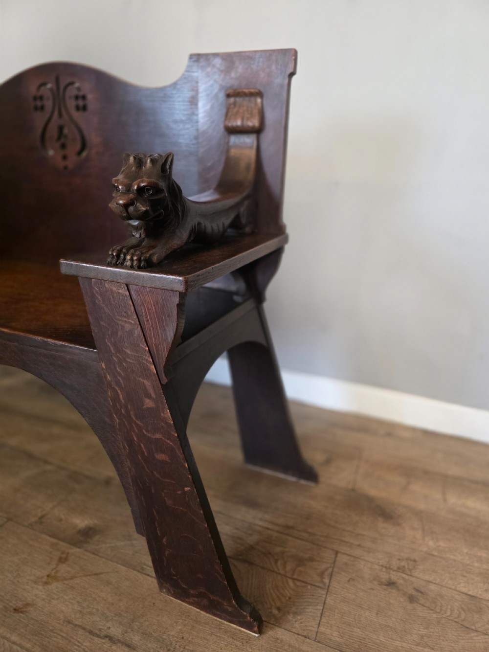 A dark oak Arts and Crafts hall bench featuring carved lion armrests, angled side legs, and a stylized inlaid motif on the backrest. View VI