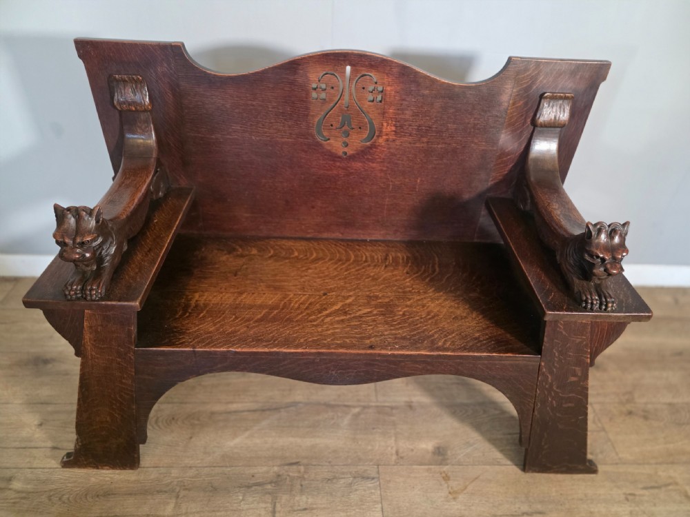 A dark oak Arts and Crafts hall bench featuring carved lion armrests, angled side legs, and a stylized inlaid motif on the backrest. View VIII