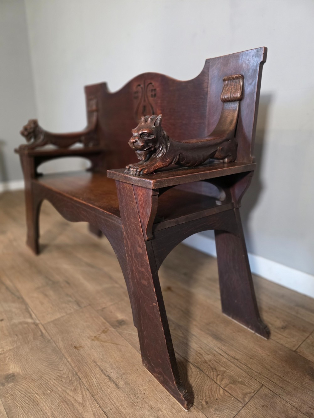 A dark oak Arts and Crafts hall bench featuring carved lion armrests, angled side legs, and a stylized inlaid motif on the backrest. View IX