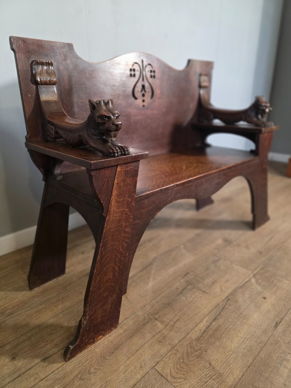 A dark oak Arts and Crafts hall bench featuring carved lion armrests, angled side legs, and a stylized inlaid motif on the backrest. View X