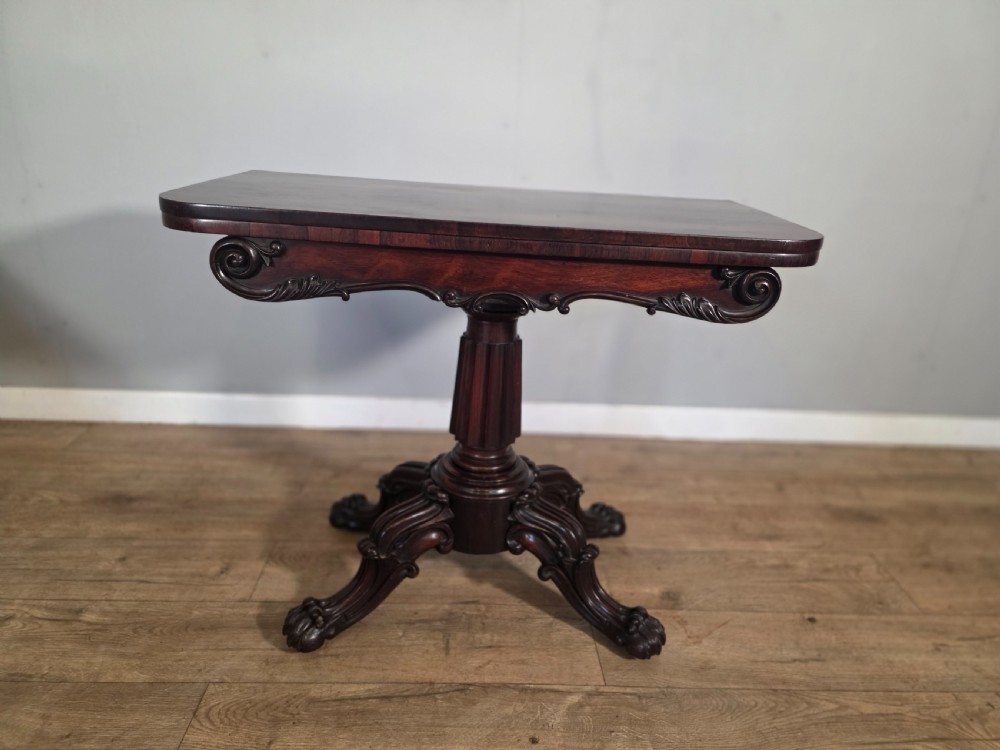 An antique early Victorian dark rosewood tea table featuring a fold-over top, carved scroll apron, and a central pedestal base with carved scroll feet.