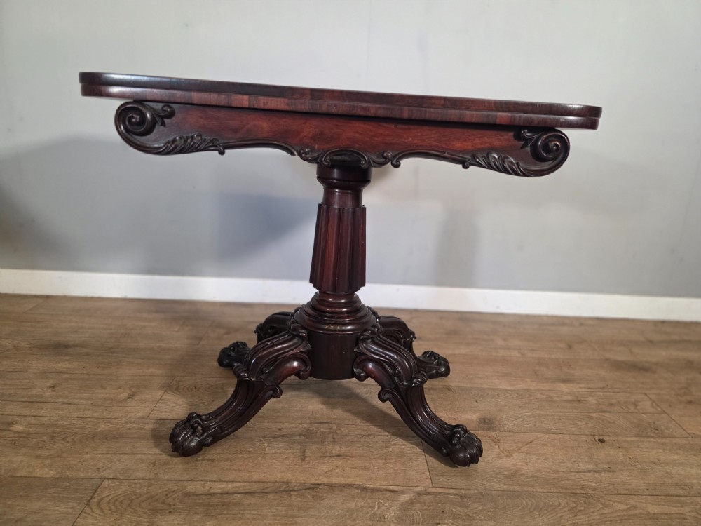 An antique early Victorian dark rosewood tea table featuring a fold-over top, carved scroll apron, and a central pedestal base with carved scroll feet. View IV