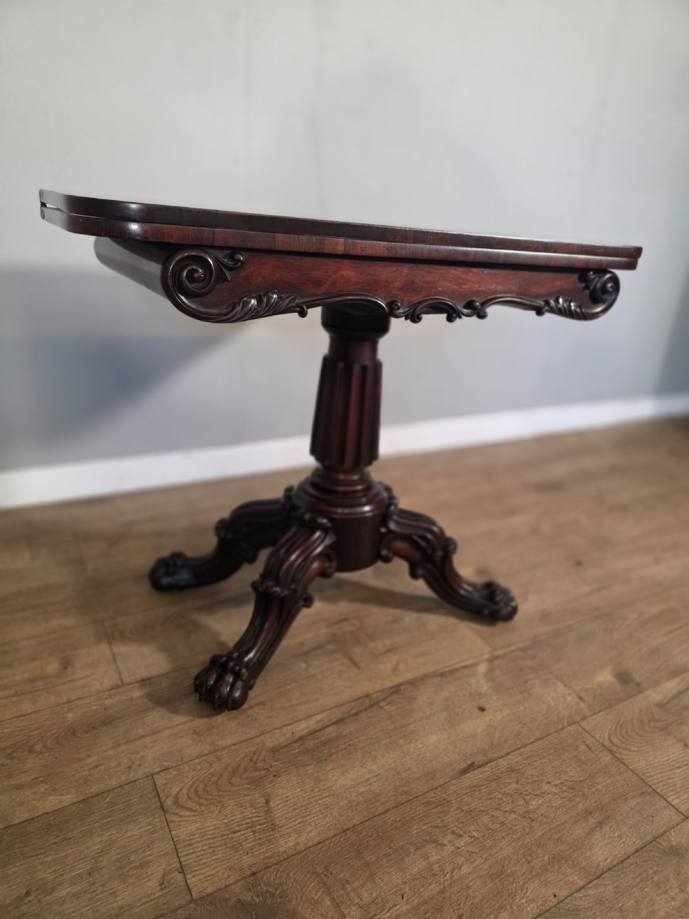 An antique early Victorian dark rosewood tea table featuring a fold-over top, carved scroll apron, and a central pedestal base with carved scroll feet. View V