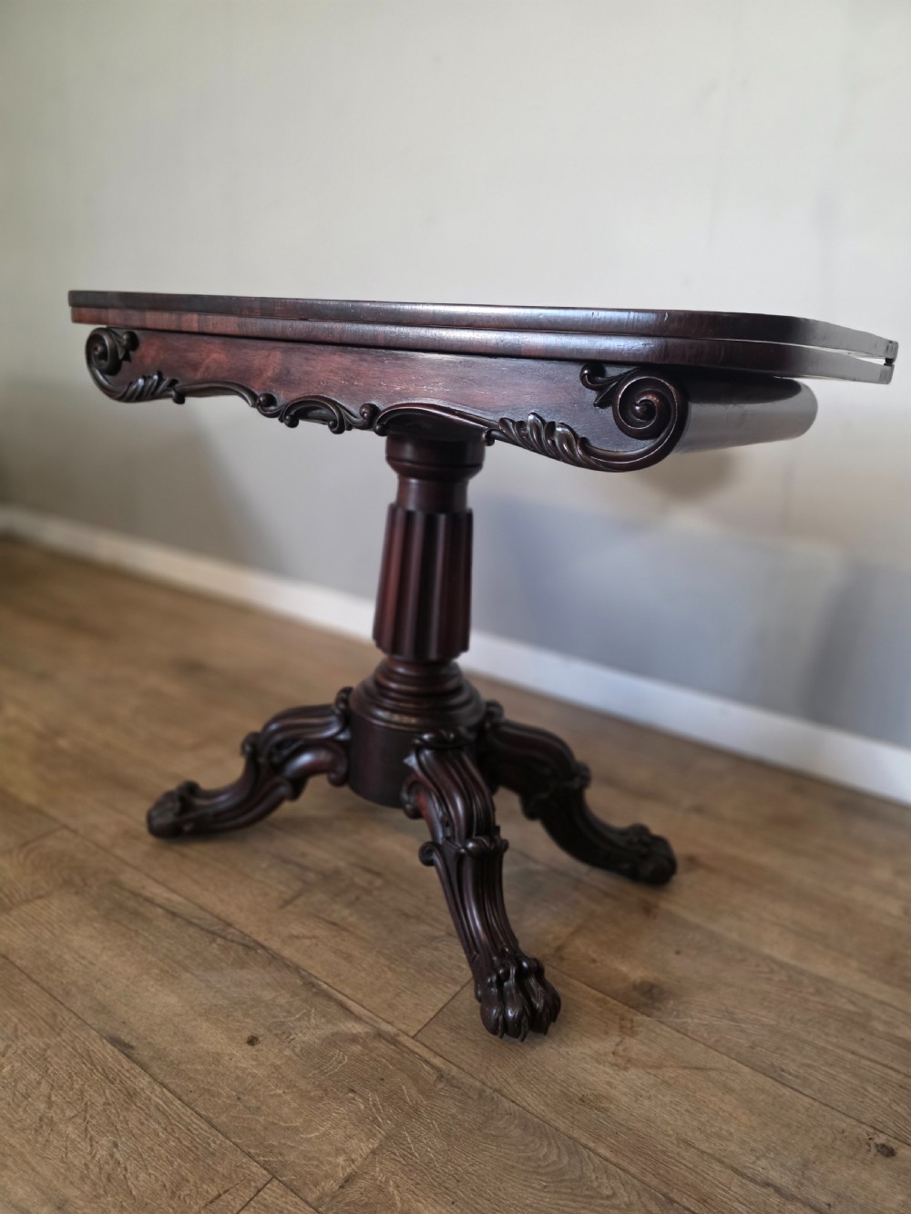 An antique early Victorian dark rosewood tea table featuring a fold-over top, carved scroll apron, and a central pedestal base with carved scroll feet. View VI