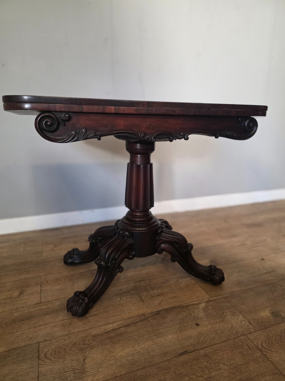 An antique early Victorian dark rosewood tea table featuring a fold-over top, carved scroll apron, and a central pedestal base with carved scroll feet. View IX