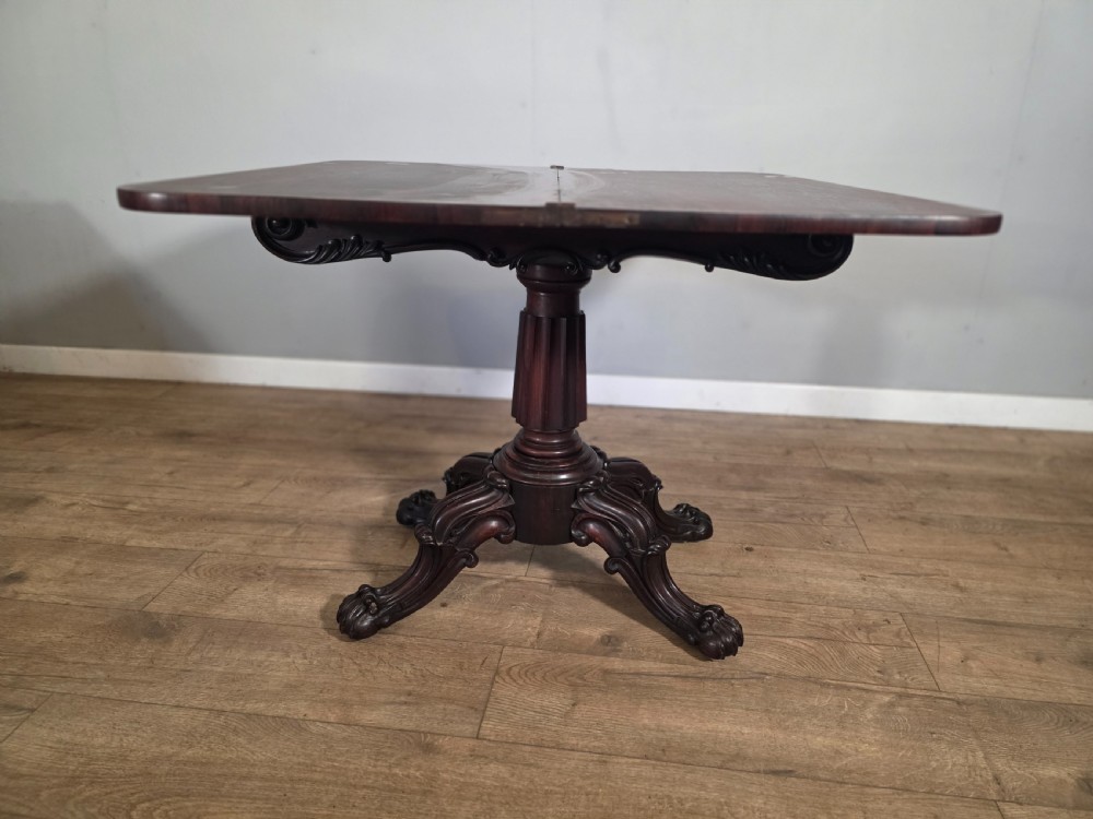 An antique early Victorian dark rosewood tea table featuring a fold-over top, carved scroll apron, and a central pedestal base with carved scroll feet. View X