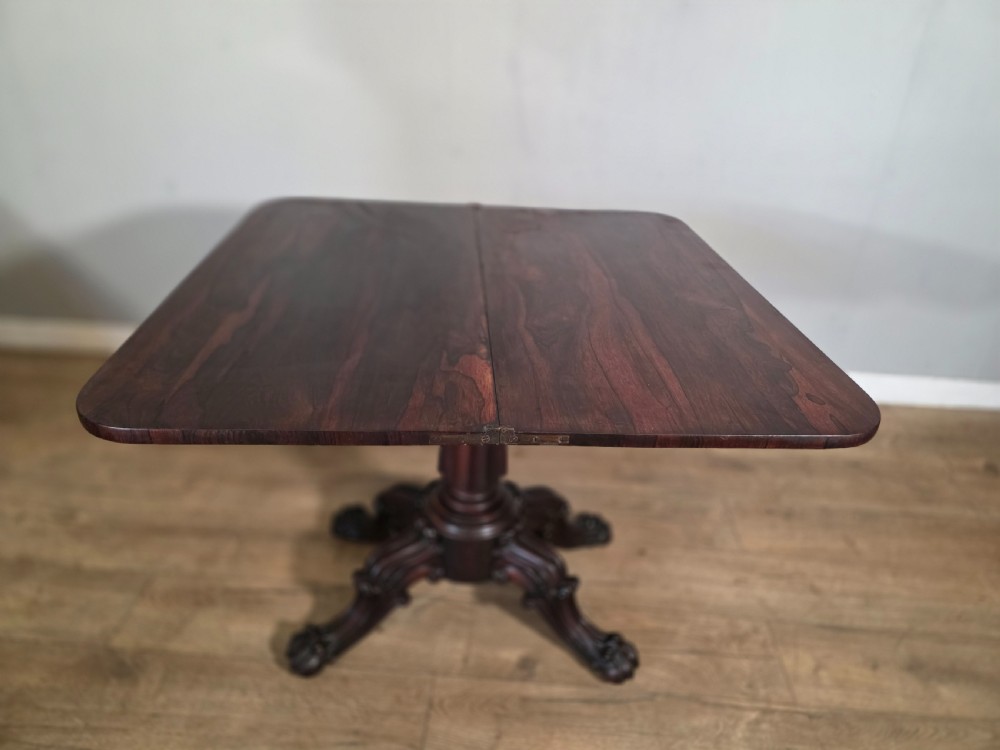 An antique early Victorian dark rosewood tea table featuring a fold-over top, carved scroll apron, and a central pedestal base with carved scroll feet. View XI
