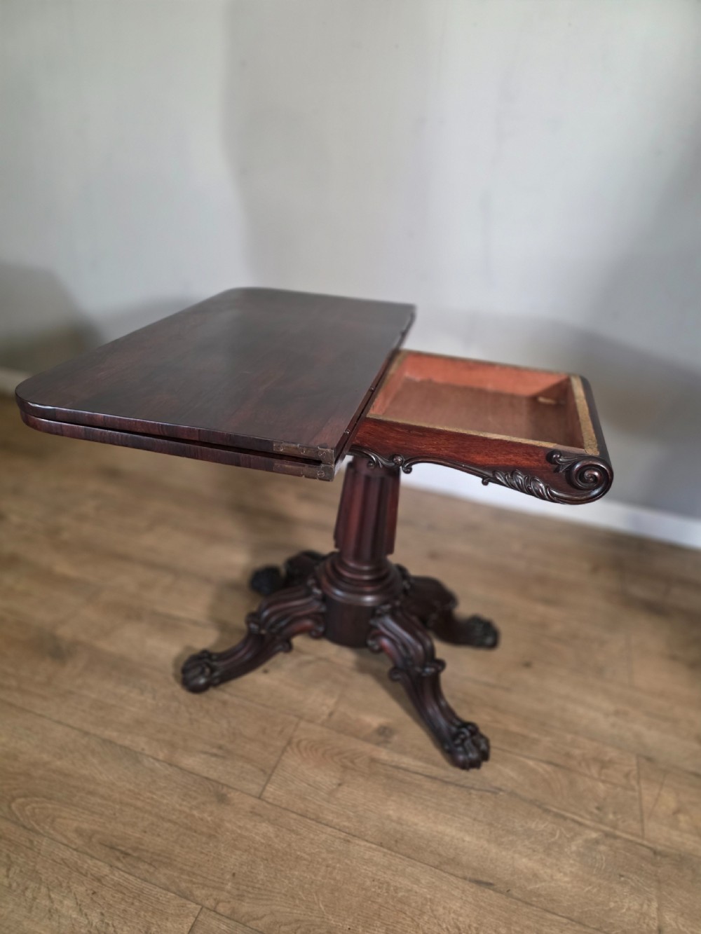 An antique early Victorian dark rosewood tea table featuring a fold-over top, carved scroll apron, and a central pedestal base with carved scroll feet. View XIV