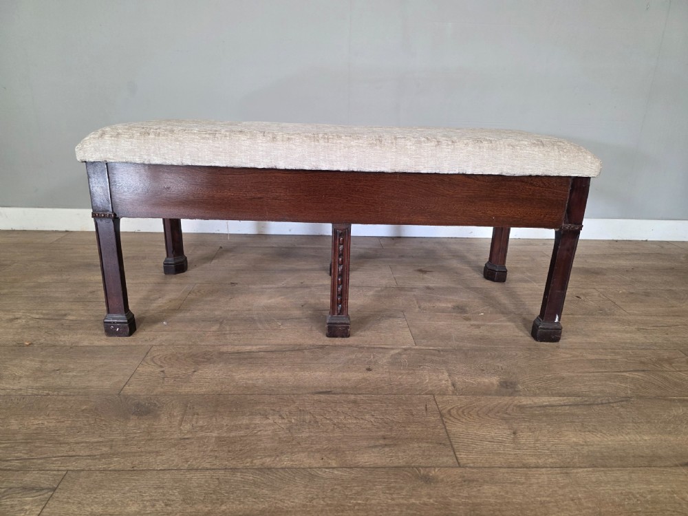 An 18th-century Adam style dark mahogany long window seat with six square tapered legs and a beige textured upholstered seat.