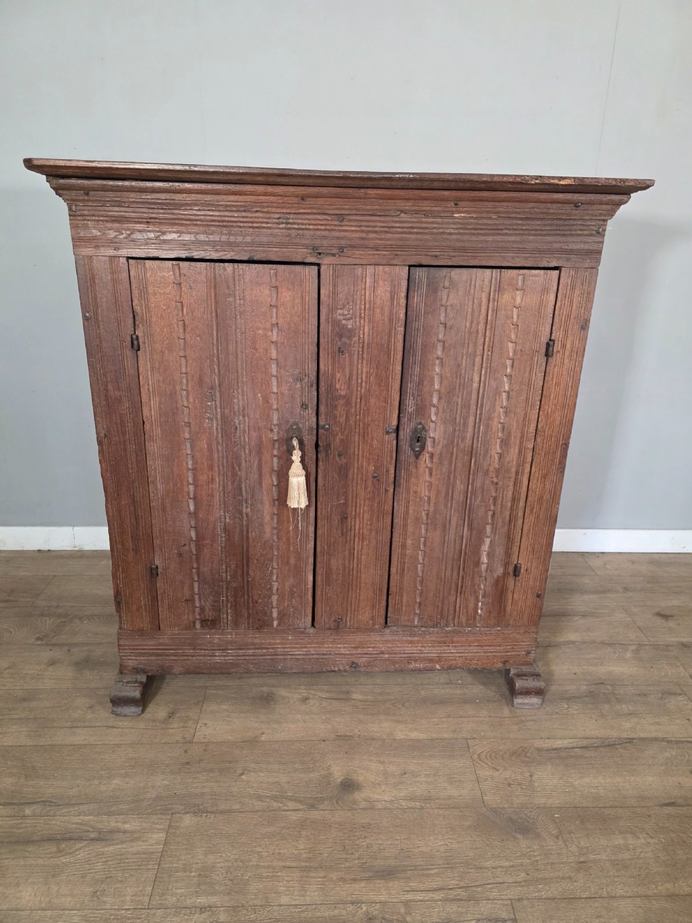 An 18th-century primitive dark oak cupboard featuring two panelled doors with notch carving, wrought iron escutcheons, and moulded top cornicing.