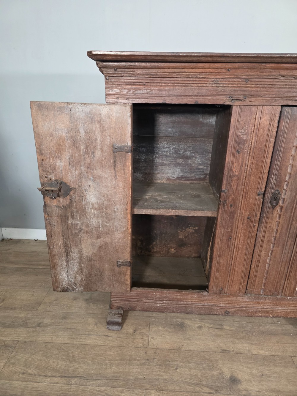 An 18th-century primitive dark oak cupboard featuring two panelled doors with notch carving, wrought iron escutcheons, and moulded top cornicing. View VII