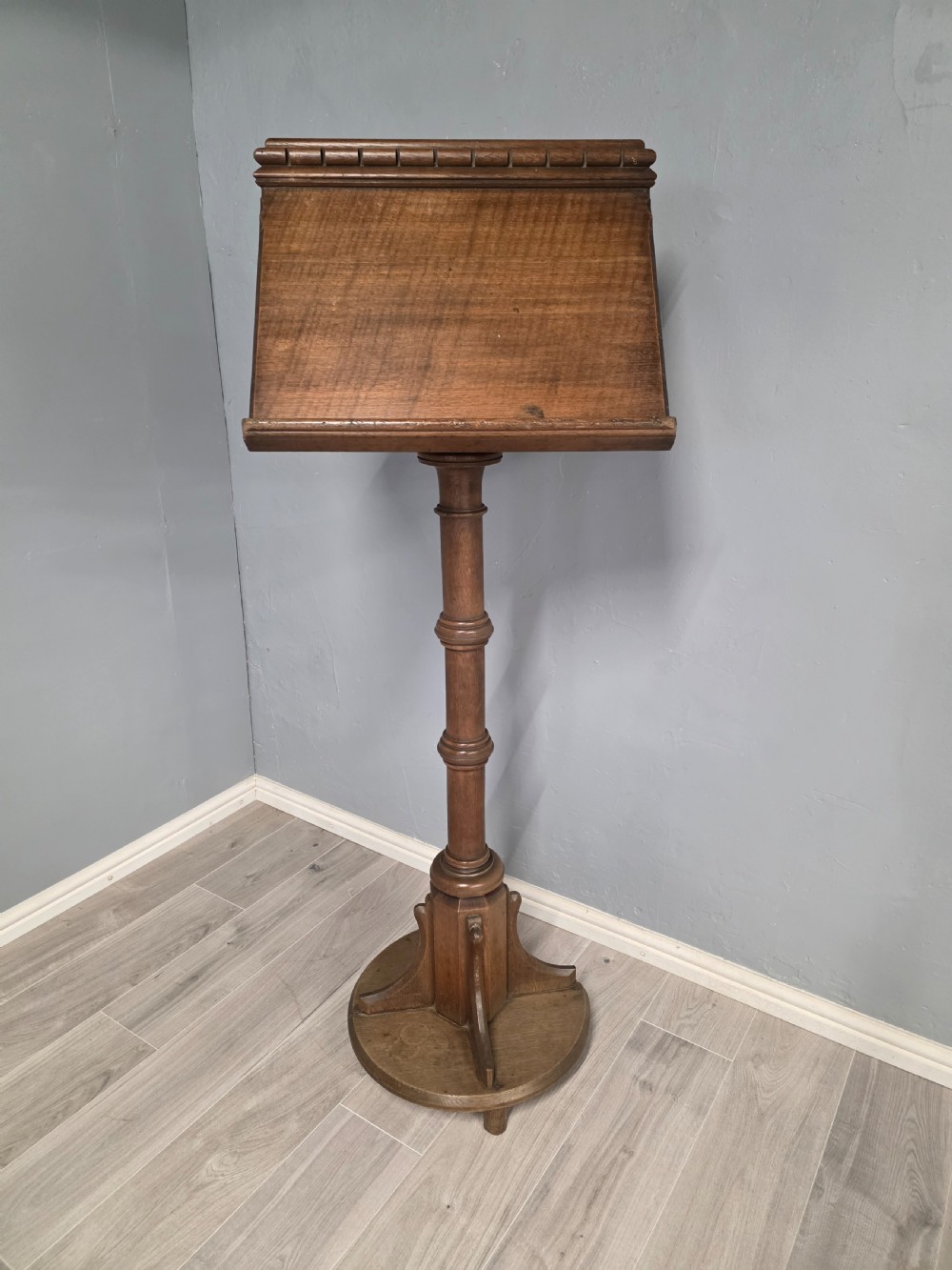 An antique Victorian oak church lectern featuring a sloped book rest with dentil moulding, a turned central column, and a circular base with bracket supports.