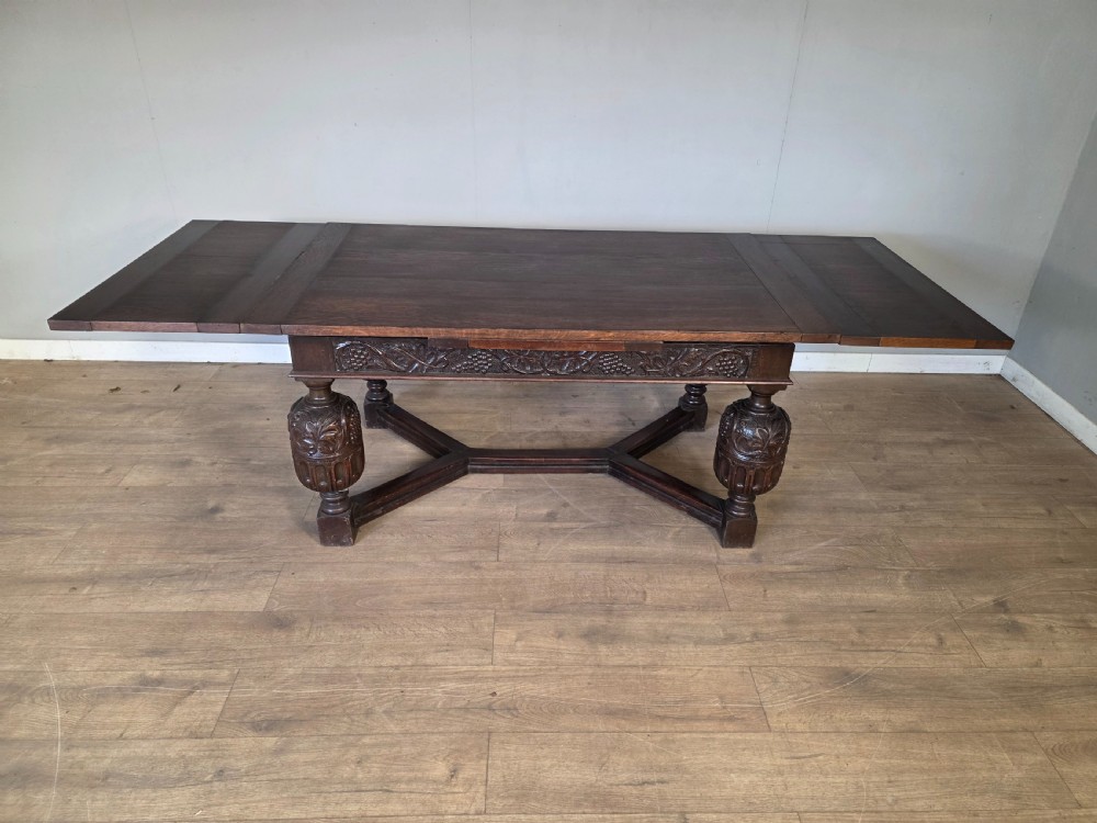 A solid dark oak extending dining table featuring carved bulbous legs, a decorative foliage apron, and a stretcher base. View X
