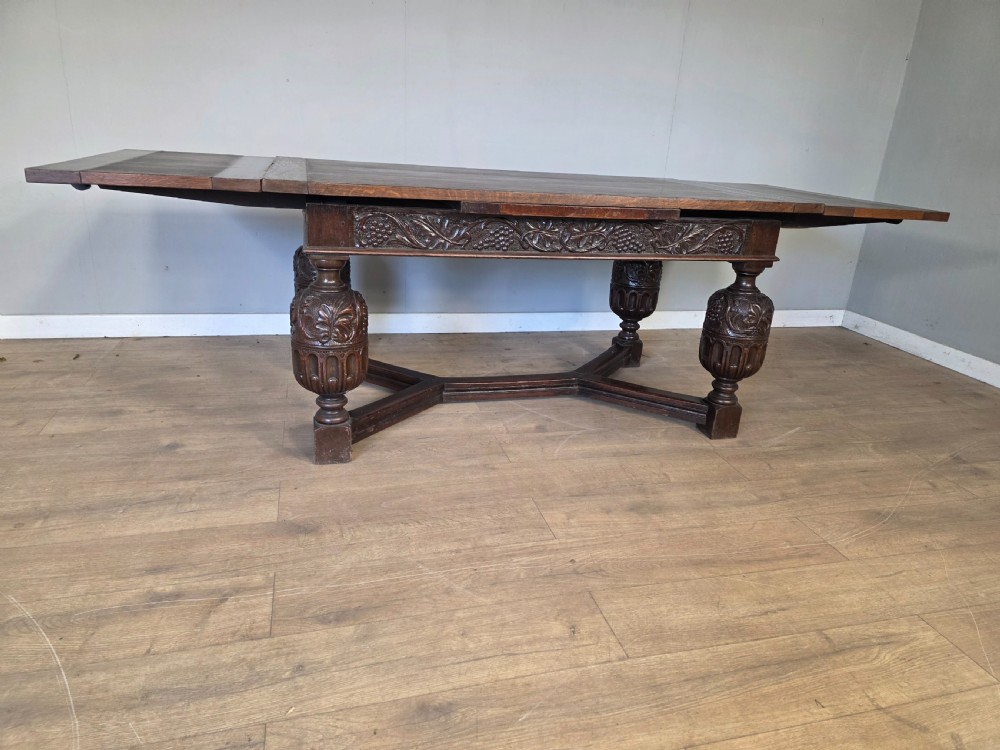 A solid dark oak extending dining table featuring carved bulbous legs, a decorative foliage apron, and a stretcher base. View XIV