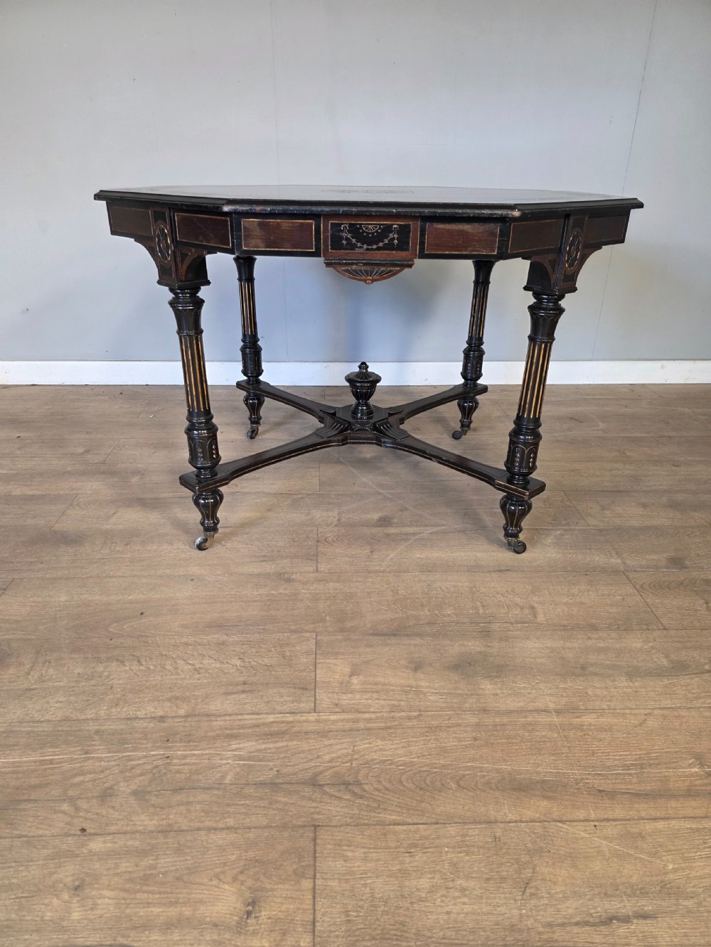 An antique Victorian octagonal centre table featuring an ebonised black finish, gilded accents, turned legs, an X-frame stretcher with a central finial, and brass castors.