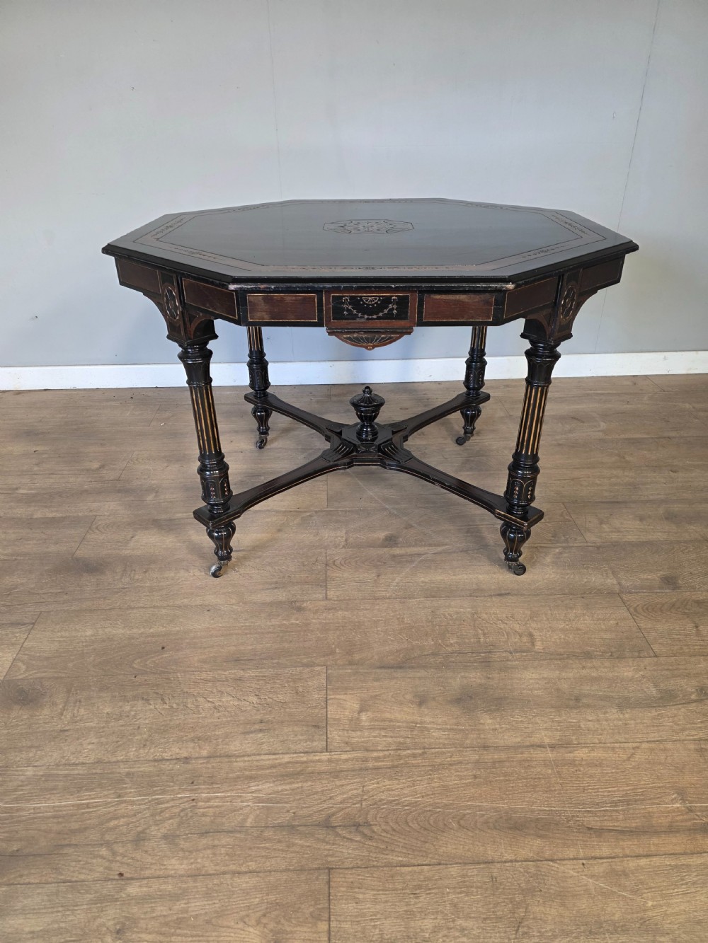 An antique Victorian octagonal centre table featuring an ebonised black finish, gilded accents, turned legs, an X-frame stretcher with a central finial, and brass castors. View II