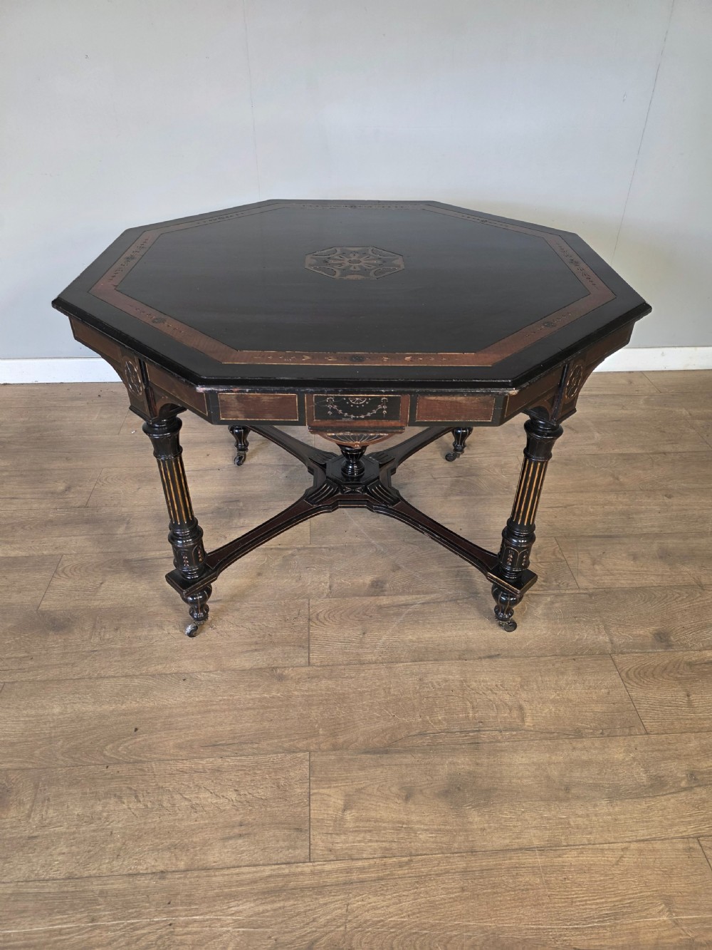 An antique Victorian octagonal centre table featuring an ebonised black finish, gilded accents, turned legs, an X-frame stretcher with a central finial, and brass castors. View III