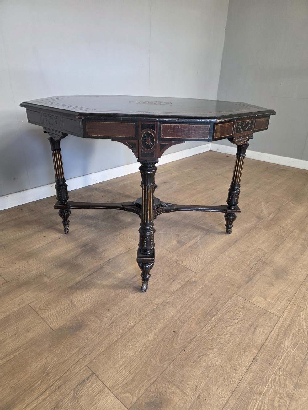 An antique Victorian octagonal centre table featuring an ebonised black finish, gilded accents, turned legs, an X-frame stretcher with a central finial, and brass castors. View IV