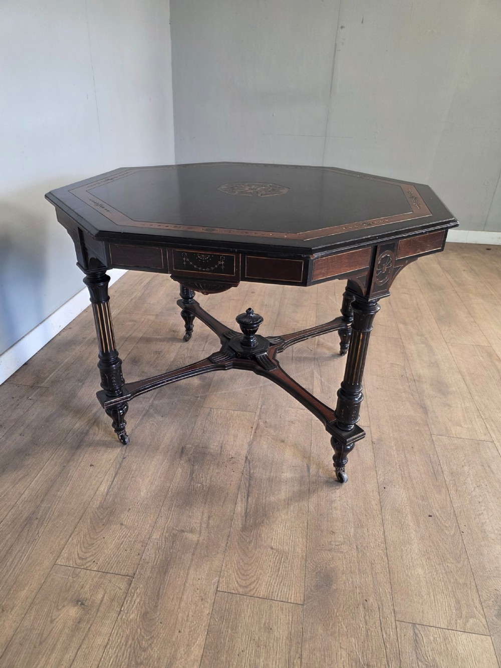 An antique Victorian octagonal centre table featuring an ebonised black finish, gilded accents, turned legs, an X-frame stretcher with a central finial, and brass castors. View V